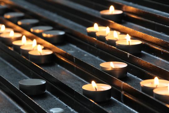 Teelichter mit flammendem Docht stehen in Reihen auf einem schwarzen Metallgestell.