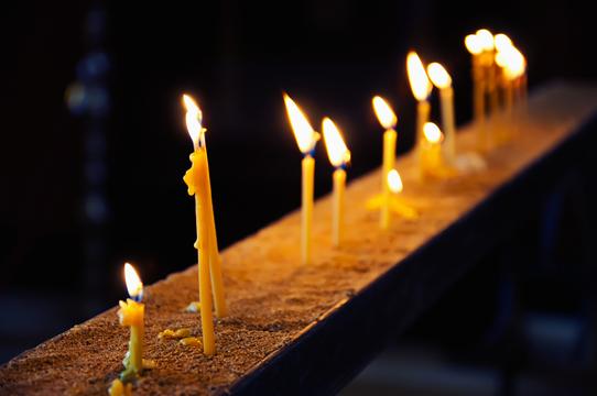 Brennende Kerzen auf einem sandigen Grund in schwachem Licht, die eine ruhige und feierliche Atmosphäre erzeugen.